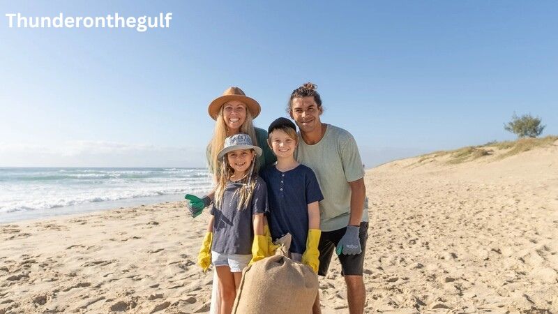 A smiling family of four stands on a sunny beach holding a large burlap sack during a cleanup, demonstrating the ThunderOnTheGulf eco friendly fishing practices and coastal conservation efforts. The group, consisting of two adults and two children in sun hats and work gloves, poses against a backdrop of rolling ocean waves and sandy dunes under a clear blue sky.