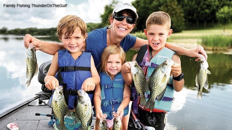 Family fishing ThunderOnTheGulf moment with a smiling adult and three children wearing life jackets on a boat, proudly holding several freshly caught fish beside a calm lake on a sunny day.